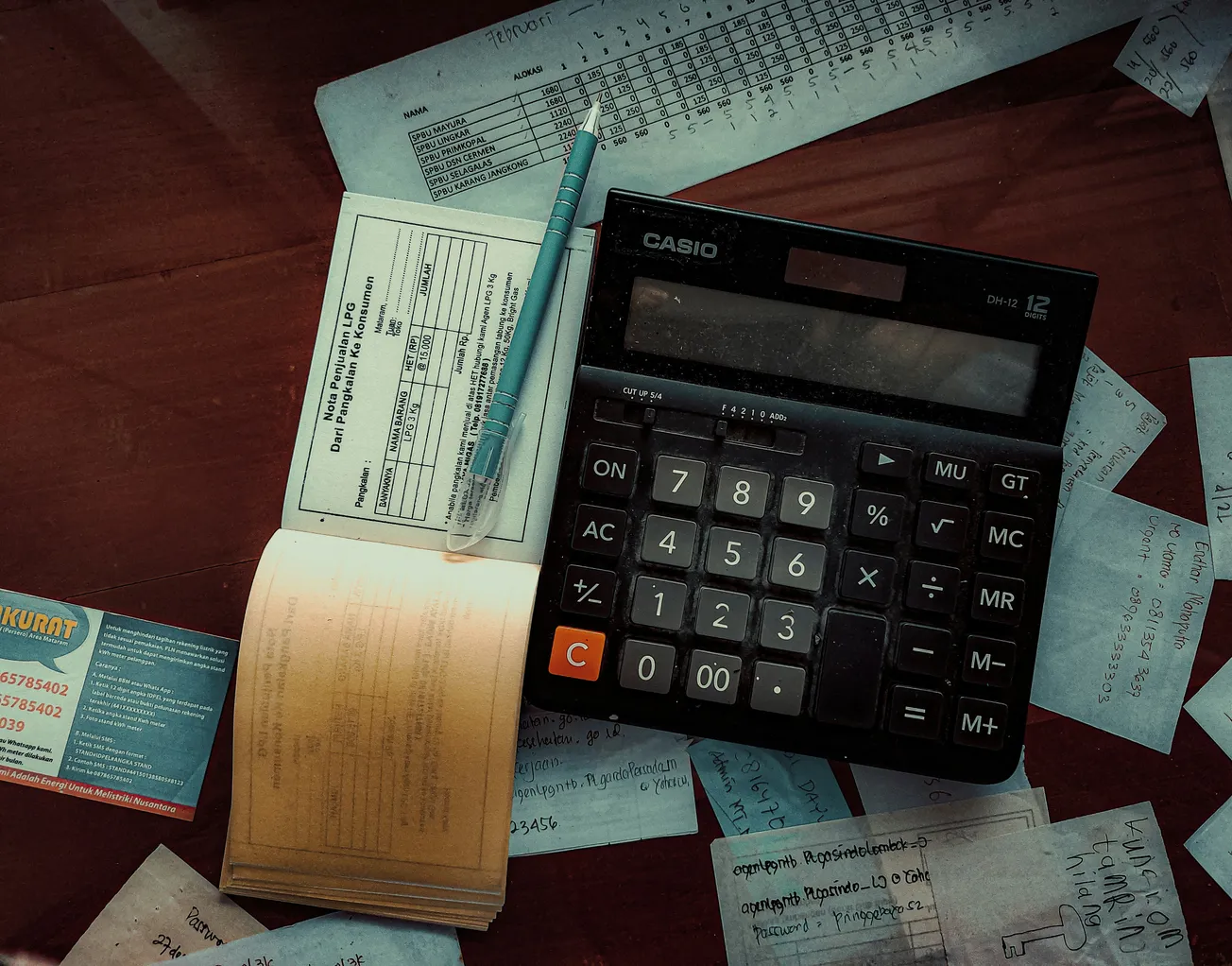 A calculator on a table surrounded by scattered papers, a pen, and forms, conveying a busy, focused atmosphere of financial calculation or bookkeeping.