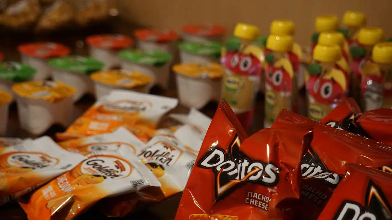 A variety of snacks on display: Doritos bags in the foreground, Goldfish crackers, applesauce cups, and colorful drink pouches in the background.