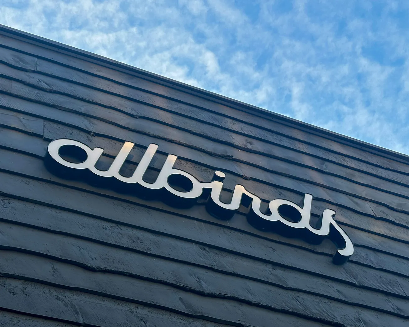 Sign for the store "Allbirds" in white, placed on a textured gray wall, with a partly cloudy blue sky in the background, conveying a fresh, modern feel.