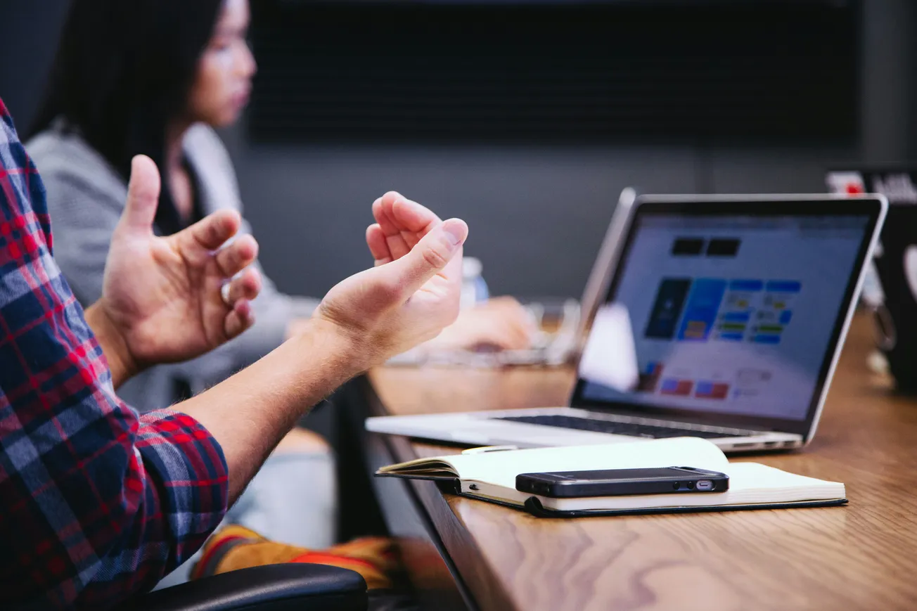 A person in a plaid shirt gestures expressively during a meeting, with a blurred colleague and laptop screen displaying a colorful presentation in the background.