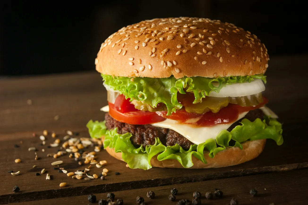 A juicy burger with lettuce, tomato, onion, pickles, cheese, and a patty in a sesame seed bun, on a rustic wooden table with scattered seeds.