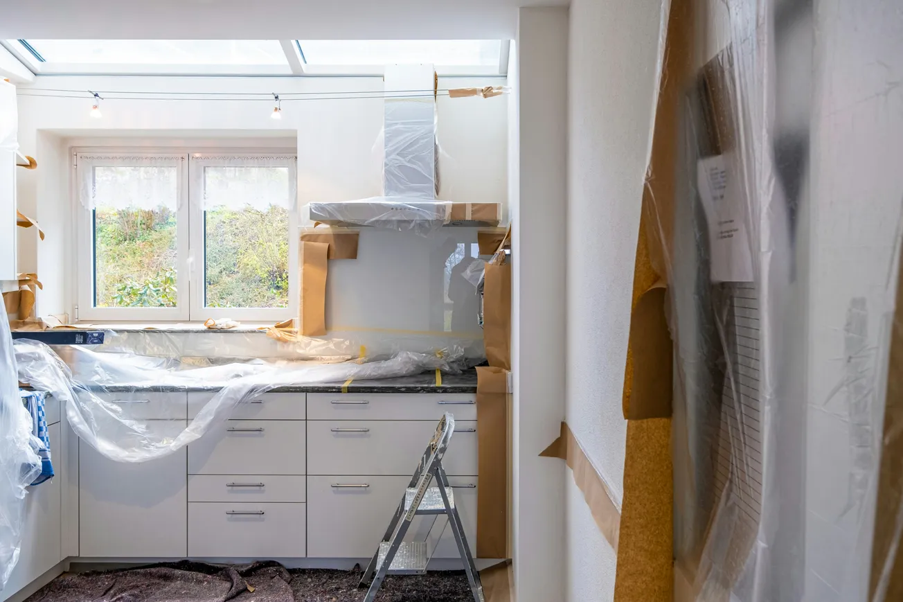 Kitchen under renovation with covered cabinets and appliances in plastic and cardboard. A step ladder stands in front of a window, creating a work-in-progress atmosphere.