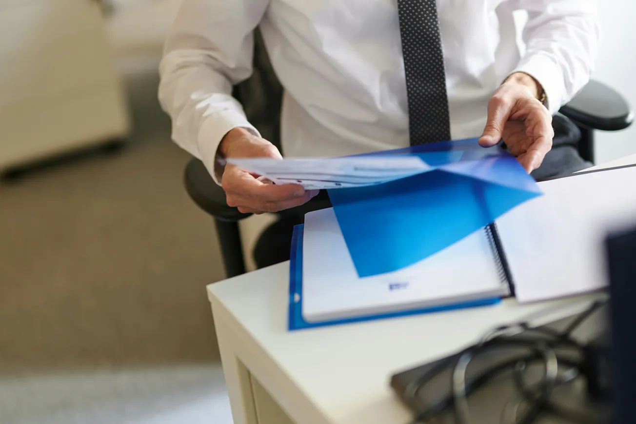 A person in a white shirt and polka-dot tie examines documents from a blue folder on a desk, conveying a professional office setting.