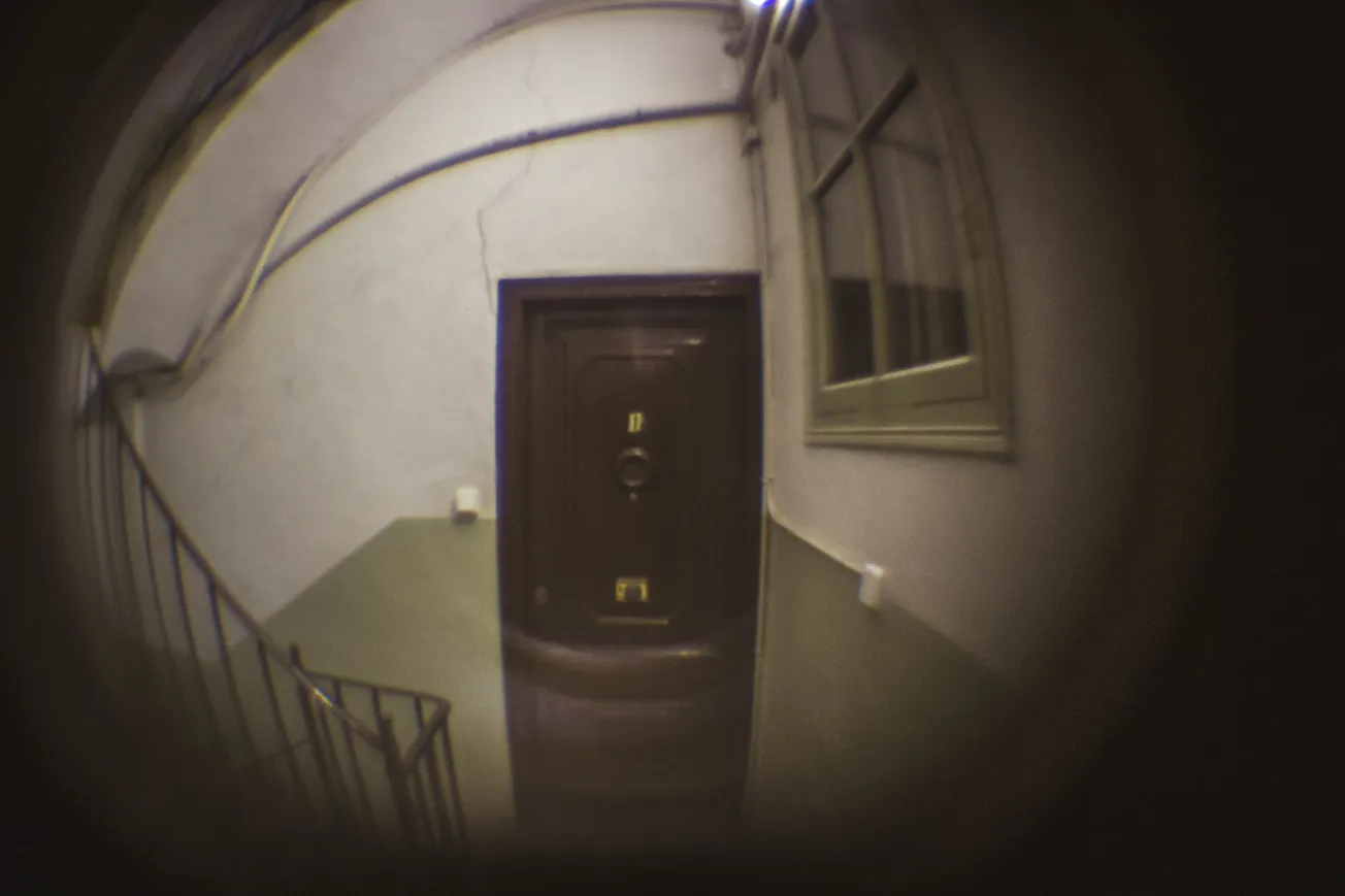 View through a fisheye lens of an old, dimly lit hallway with a closed wooden door, metal railing on the left, creating a mysterious, eerie tone.