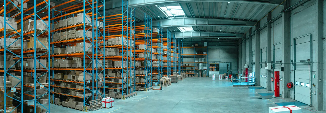 A spacious, well-lit warehouse interior with high shelves filled with boxes. The floor is clear. Emergency equipment lines one wall, conveying order and readiness.