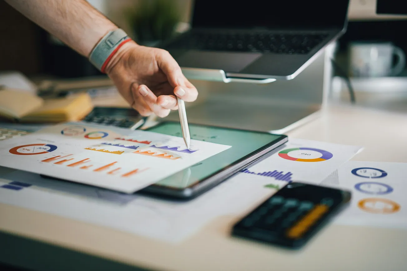 A person's hand uses a pen to point at a colorful bar chart on a paper over a tablet. A laptop and financial documents are visible on the desk.