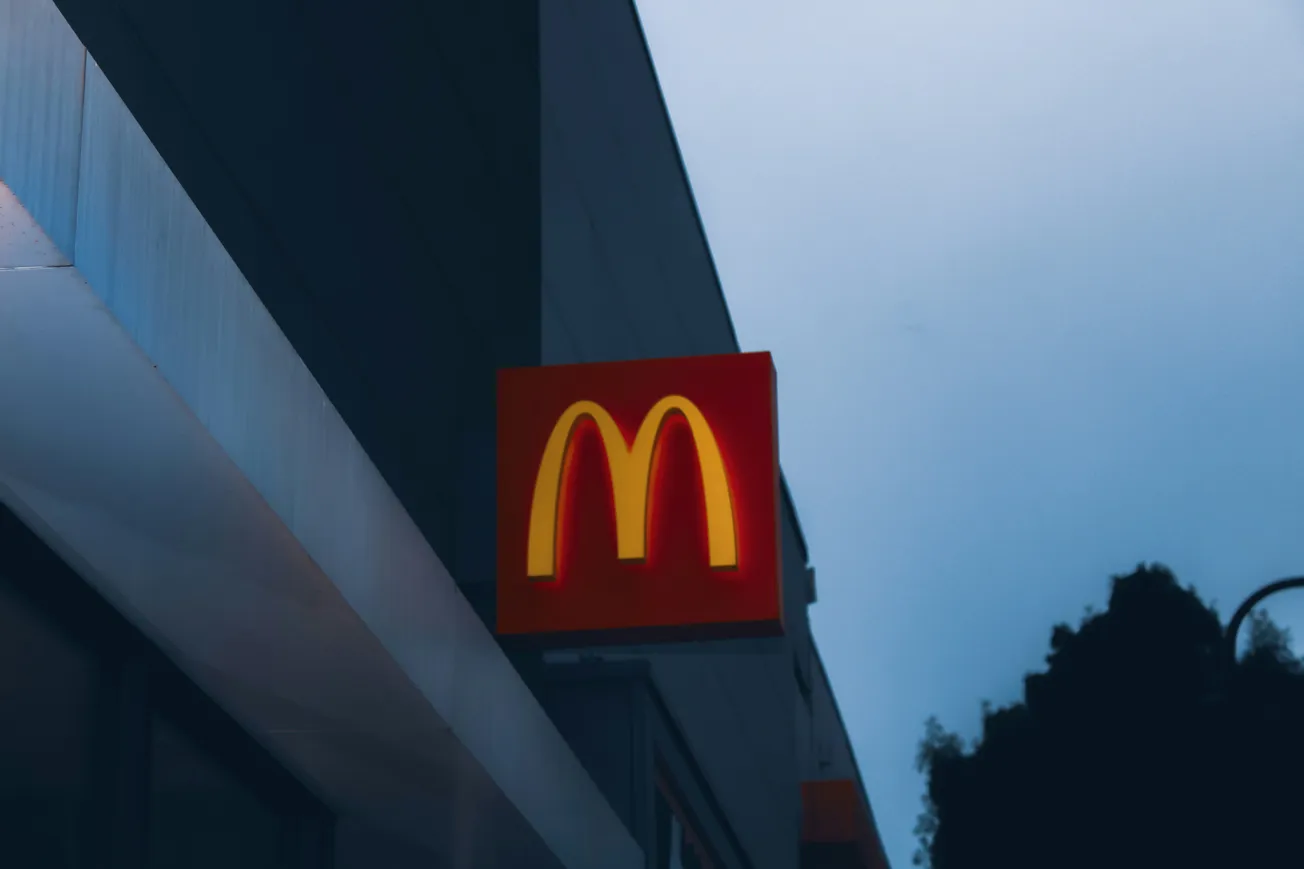 The image shows a brightly lit McDonald's logo against a darkening sky, conveying a sense of evening. The red and yellow glow adds warmth and familiarity.