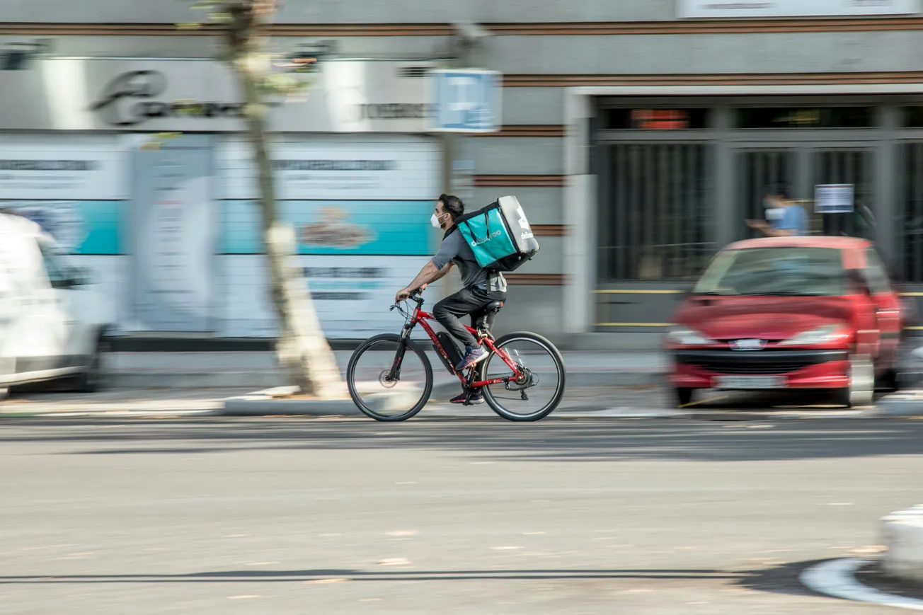 A delivery cyclist rides swiftly down an urban street, wearing a mask and a teal delivery backpack. Motion blur highlights the fast pace.
