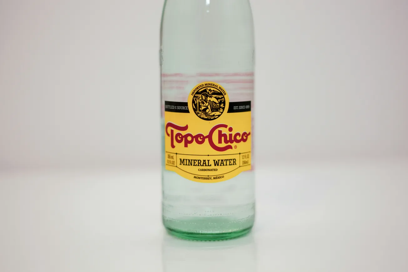 Clear glass bottle of Topo Chico mineral water on a white background. The label is yellow with red text and a black emblem, conveying a classic, refreshing tone.