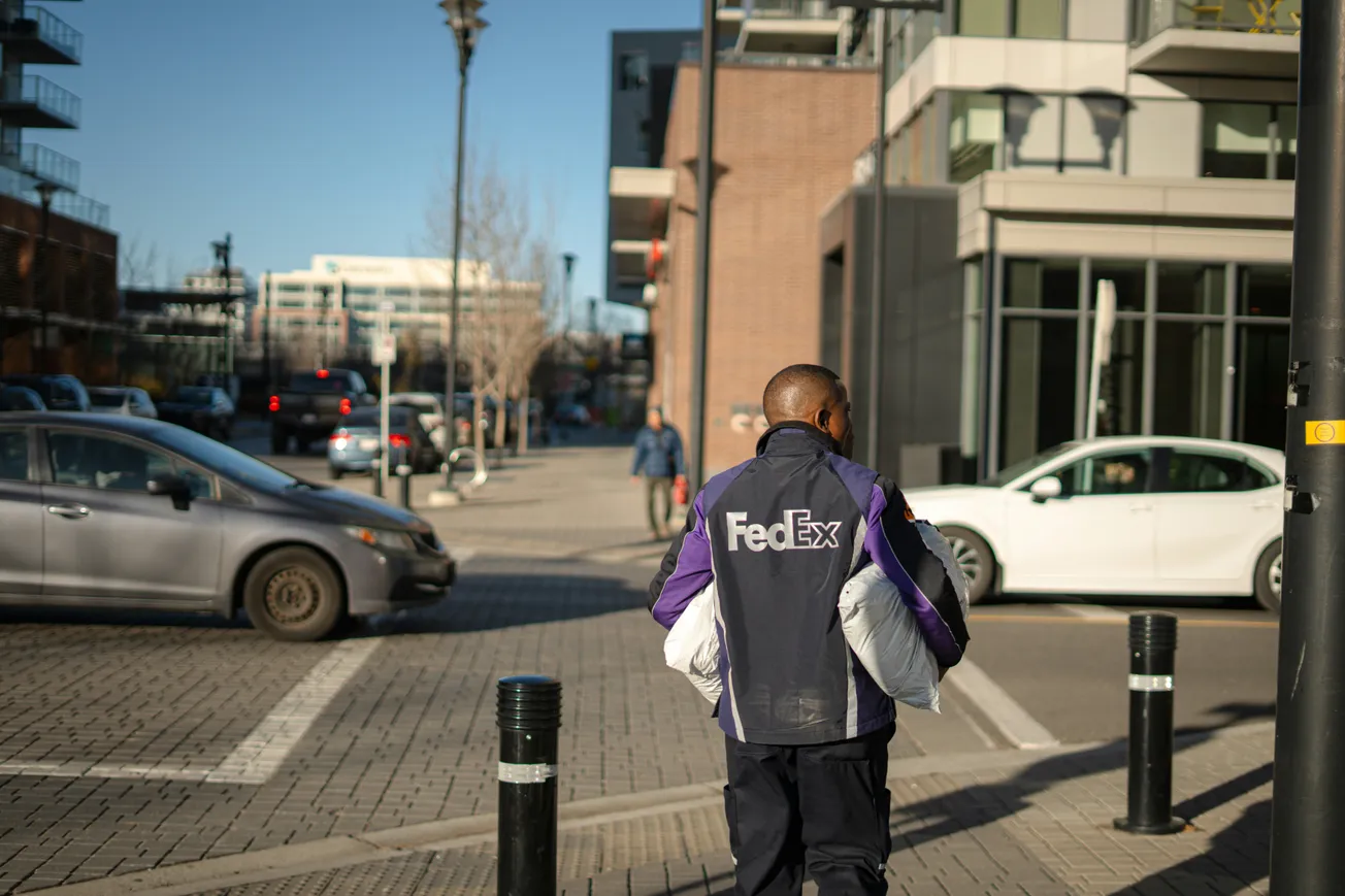A FedEx worker in uniform walks through a sunny urban area carrying packages. Cars and pedestrians are visible near modern buildings.