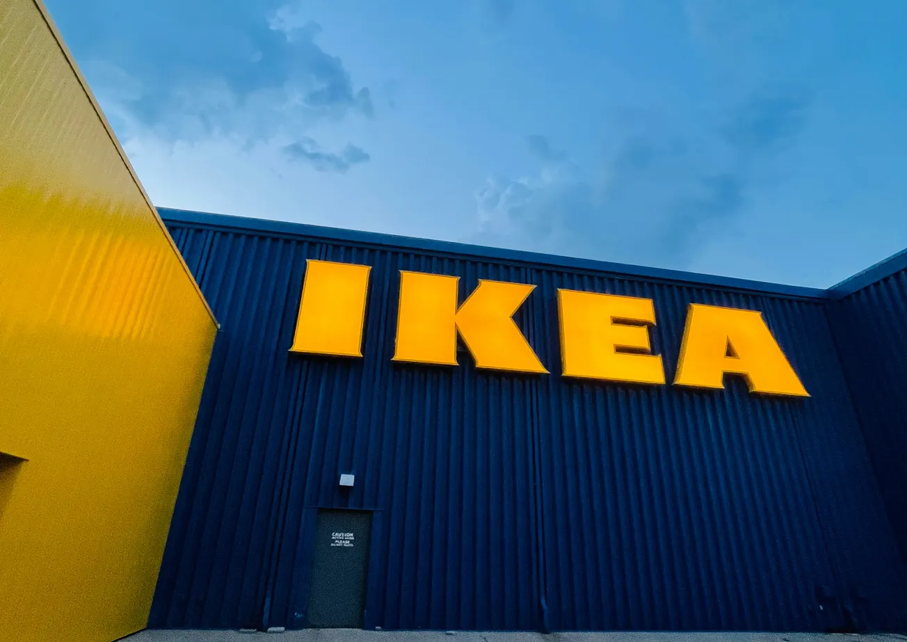 A large IKEA logo in bold yellow letters is displayed on a dark blue building, under a partly cloudy sky. The scene conveys a modern, welcoming vibe.