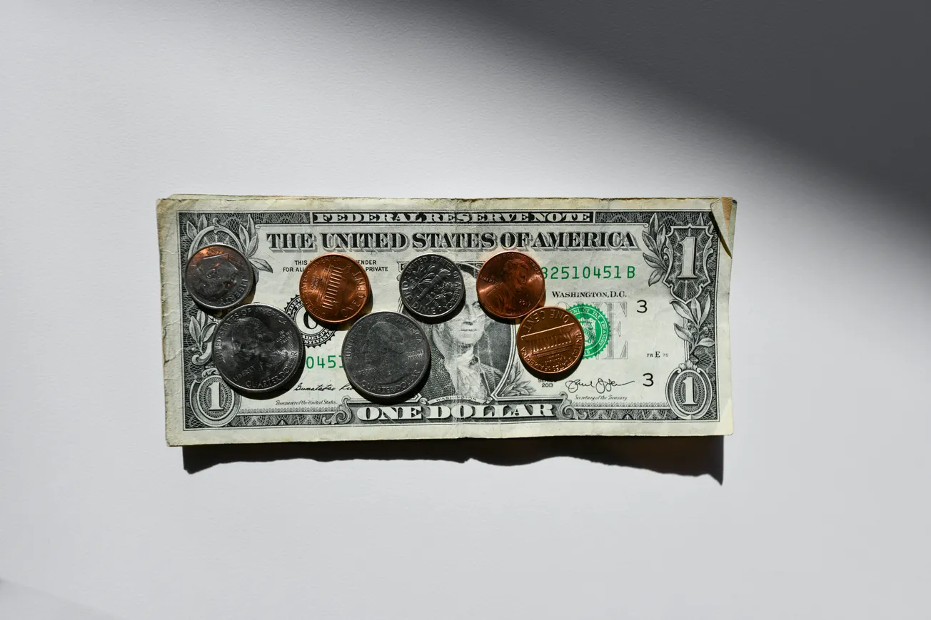 A U.S. dollar bill lies on a white surface with eight coins placed on top: a mix of pennies, nickels, dimes, and a quarter, casting shadows and creating contrast.