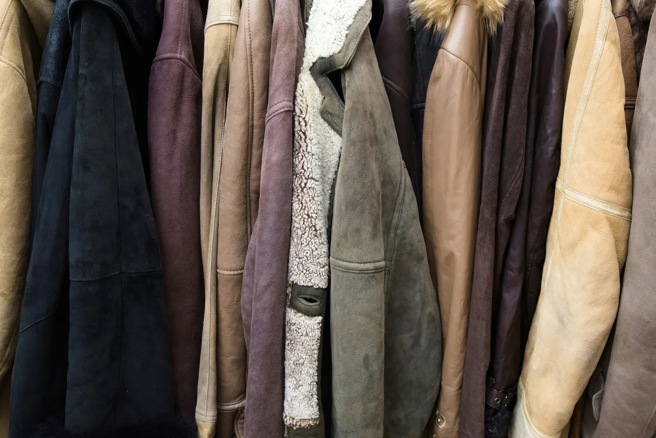 A row of various leather and suede jackets in earthy tones hangs closely together, showcasing textures like fur collars and shearling linings, evoking warmth.