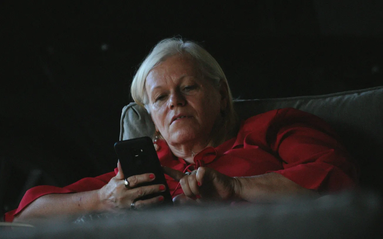 An older woman with white hair, wearing a red blouse, sits on a gray couch intently looking at her smartphone in dim lighting, creating a contemplative mood.