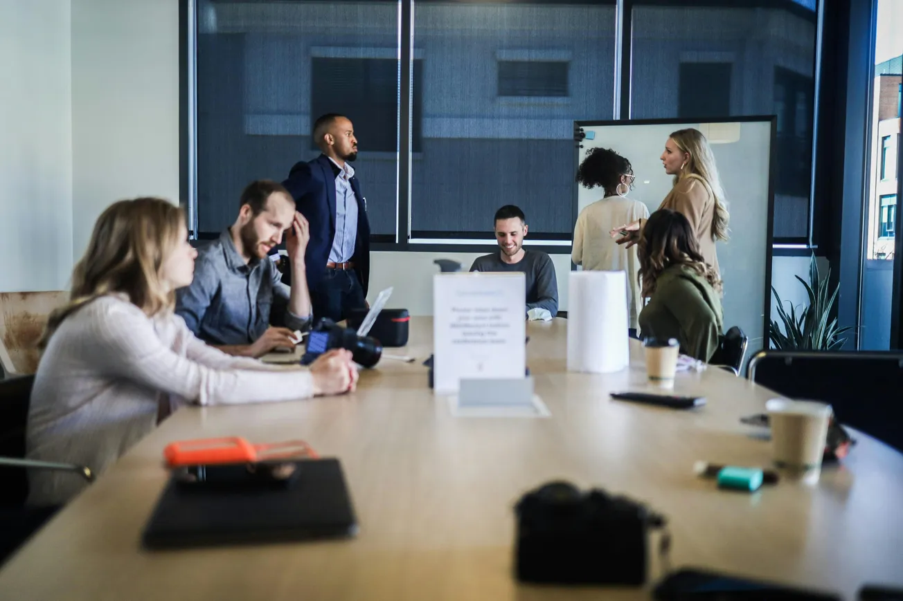 A diverse group of people in an office is having a meeting. Some are seated at a table with laptops, others are having a standing discussion by a whiteboard.