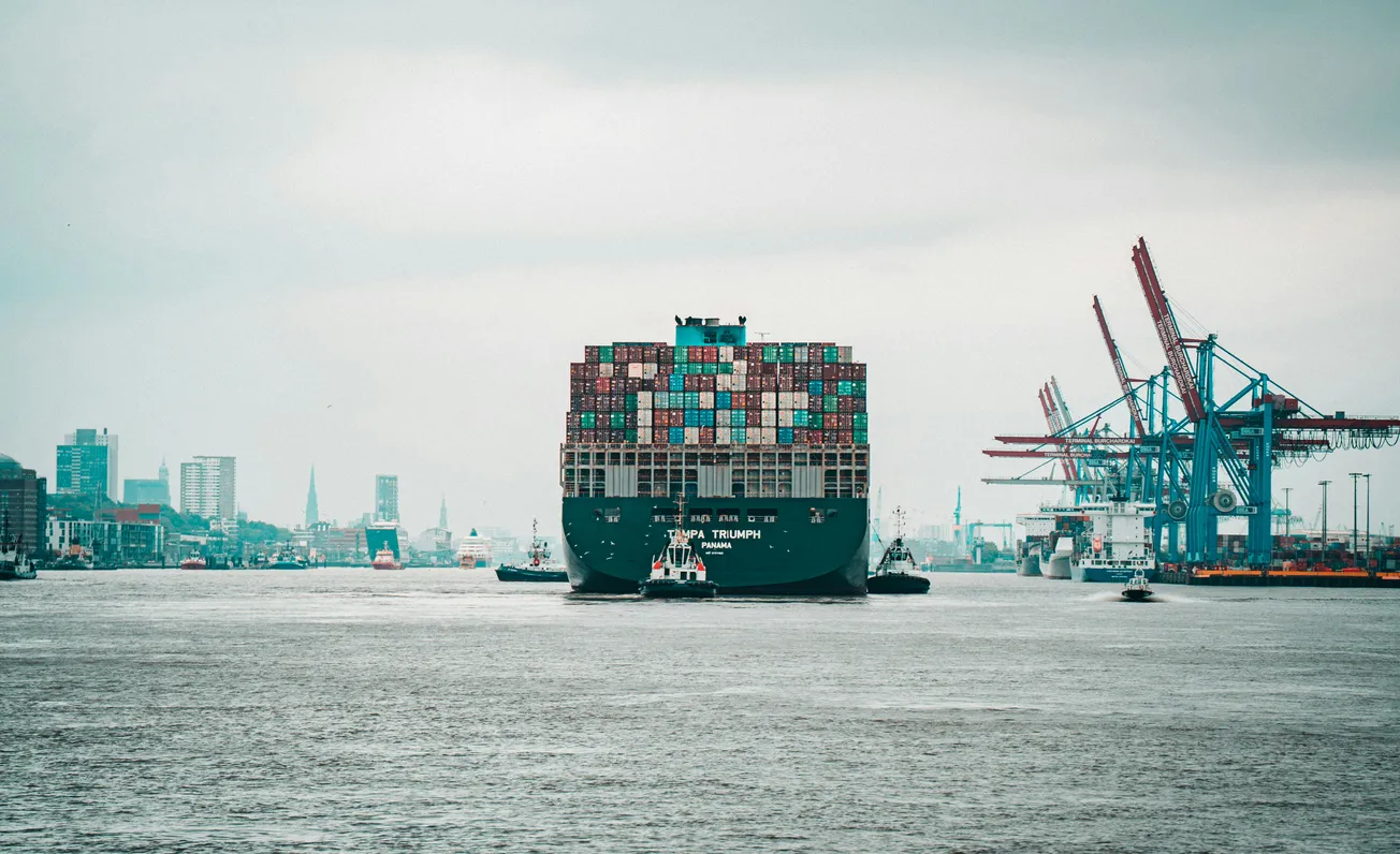A massive container ship sails in a busy port, stacked with colorful containers. Cranes line the dockside under a cloudy sky, conveying industry and scale.