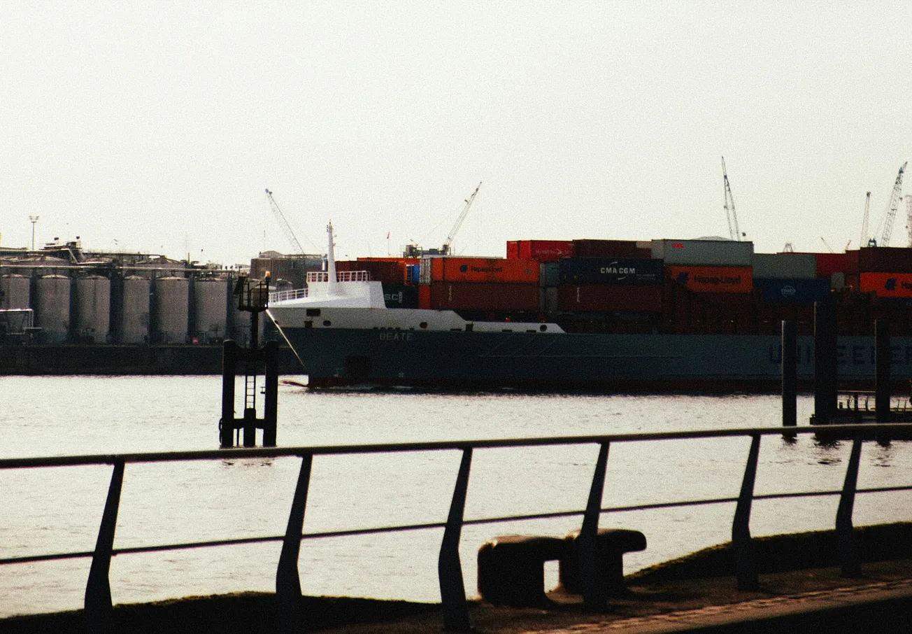 A cargo ship carrying colorful containers is docked in an industrial port. Silos and cranes are visible in the background under a cloudy sky. Calm, industrial scene.