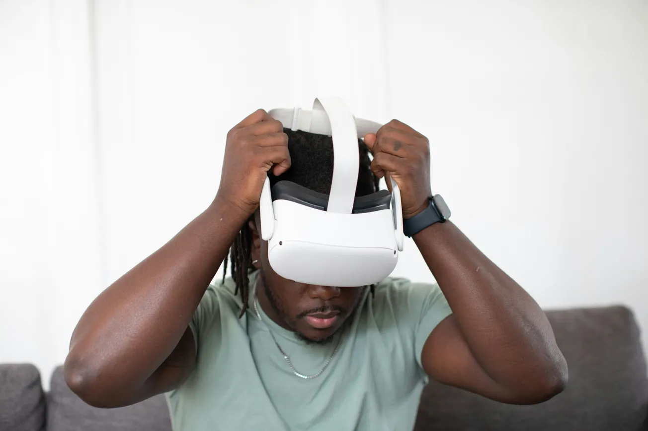 A person in a light green shirt puts on a white VR headset, sitting on a gray couch. The scene conveys focus and anticipation in a casual setting.