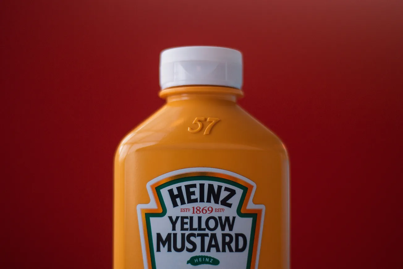 Bottle of Heinz Yellow Mustard with a white cap, shown against a deep red background. The mustard bottle is vibrant and eye-catching.