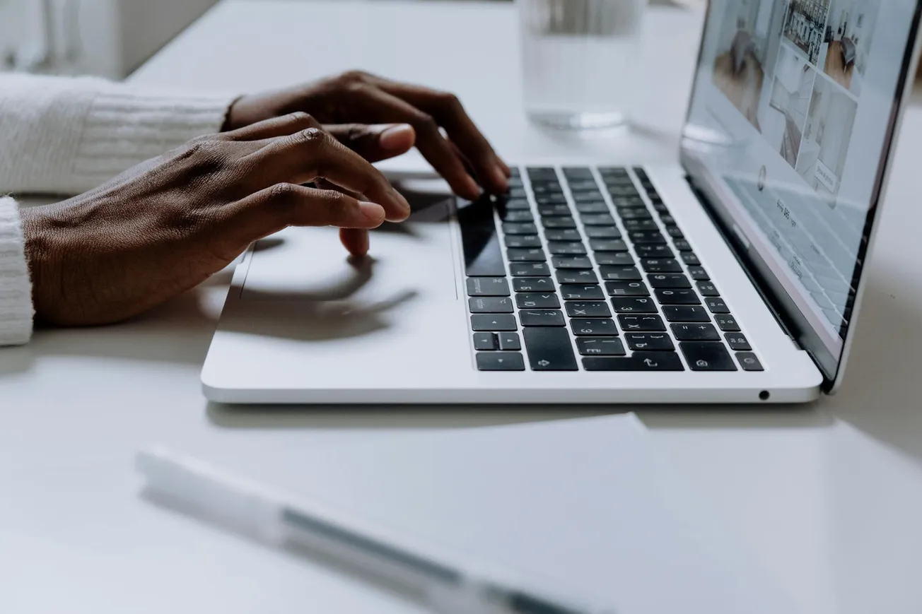 Hands typing on a laptop keyboard in a bright, minimalist setting. A white pen and glass of water are on the desk, conveying a calm, focused mood.