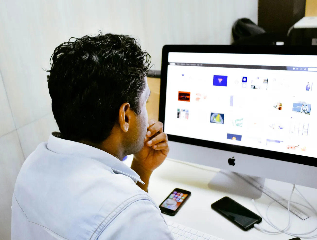 A man in a light blue shirt is focused on a computer screen displaying various images. Beside the computer are a smartphone and a tablet on the desk.