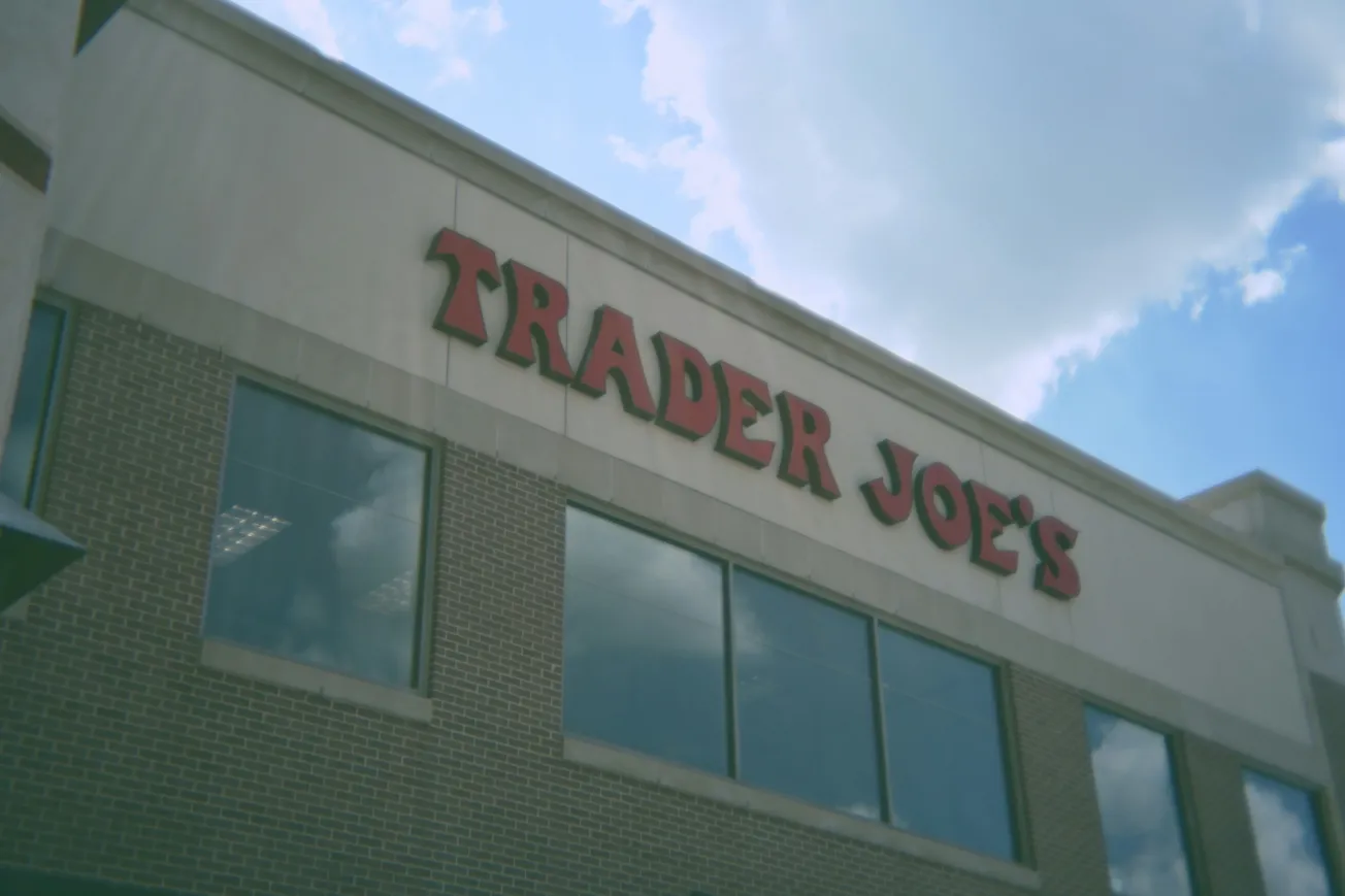 Brick building facade with large "Trader Joe's" sign in red letters. Partly cloudy sky reflects in the windows, conveying a bright, clear day.