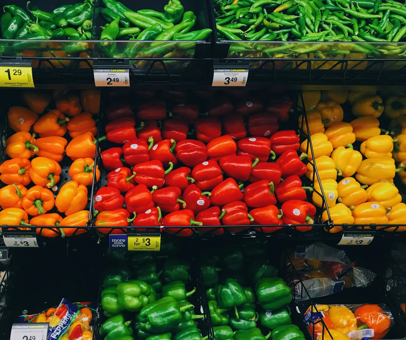 Variety of colorful bell peppers and jalapeños on display in a grocery store. Prices and signs visible; vibrant green, red, yellow, and orange tones.