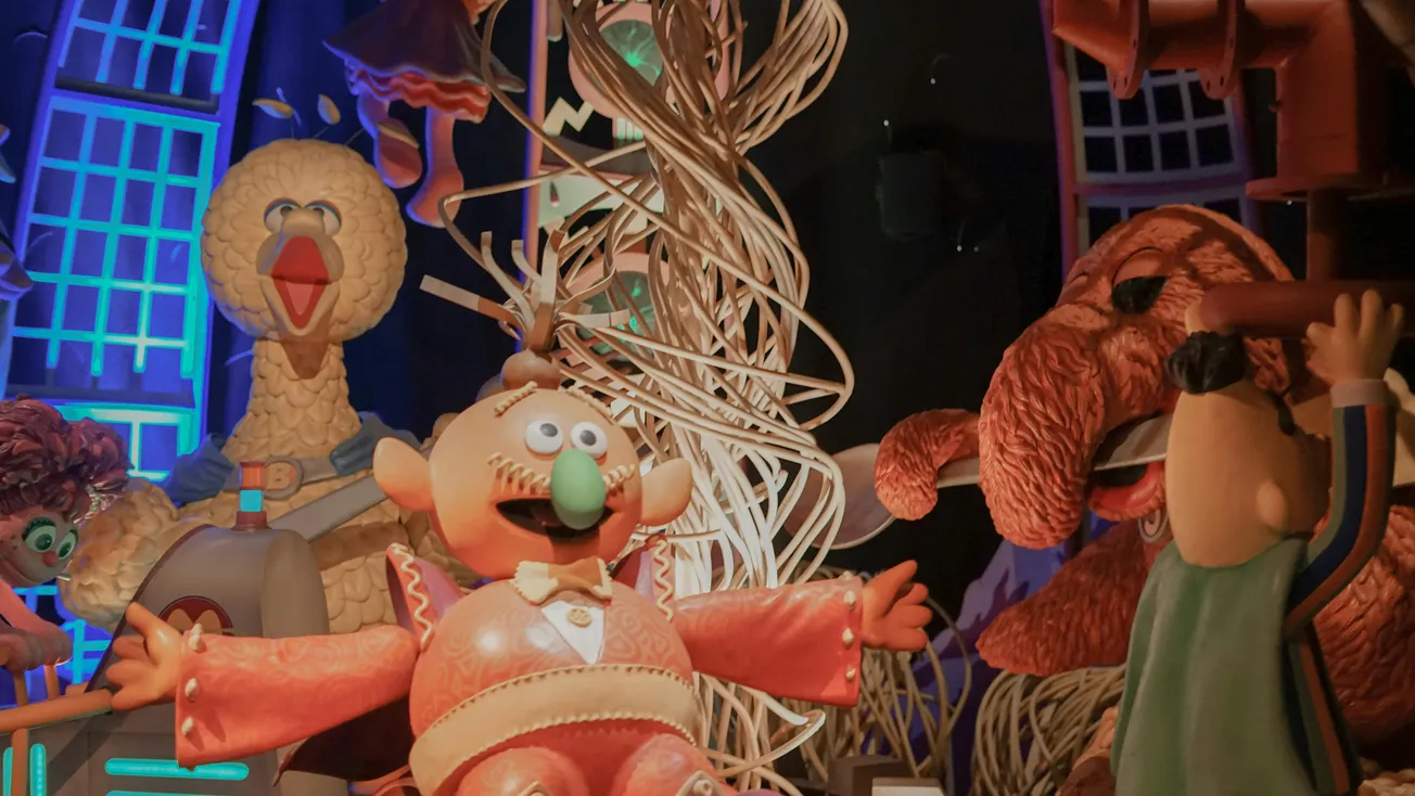 A whimsical scene with colorful, cartoonish characters in a festive setting. Includes a large yellow bird, playful decorations, and vibrant lighting.