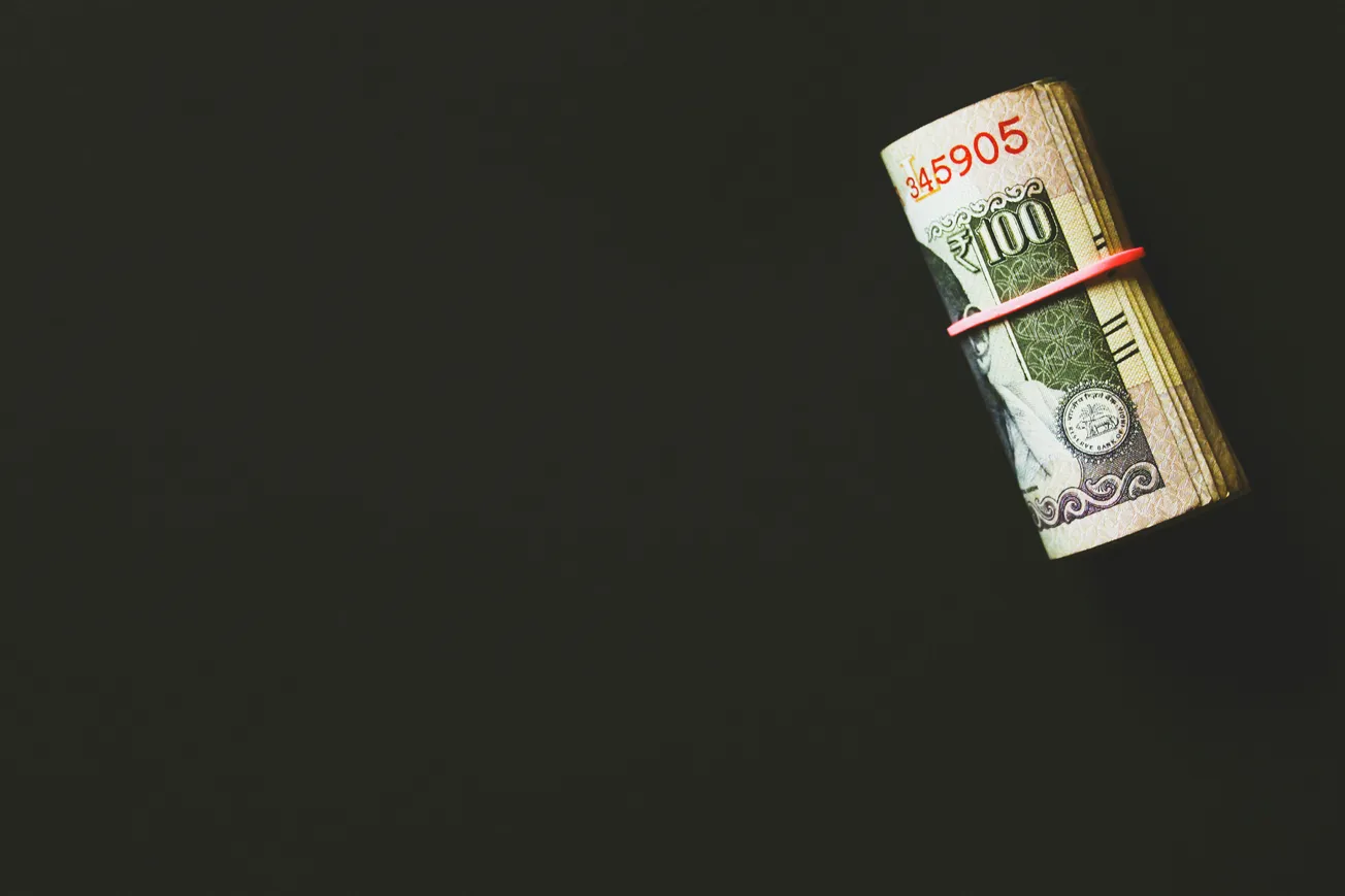 A roll of banknotes bound by a red rubber band against a black background. The tone is minimalist, highlighting themes of finance and wealth.