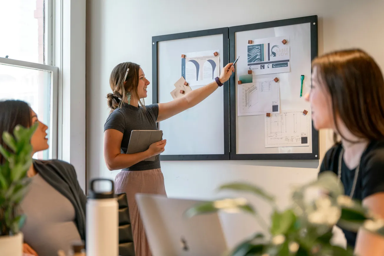 A woman gestures at charts on a bulletin board during a meeting, engaging colleagues seated around a table. The scene conveys collaboration and focus.