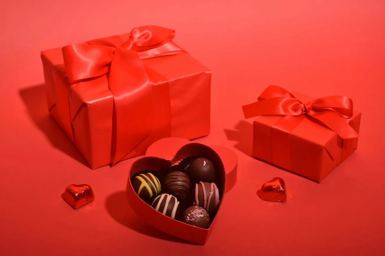 Red background with two gift boxes wrapped in red paper and bows. A heart-shaped box of assorted chocolates is open, surrounded by two foil hearts.