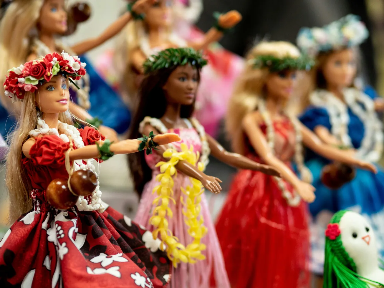 Dolls dressed in colorful Hawaiian attire, featuring leis and floral headbands, are posed as if dancing hula. The scene is vibrant and festive.