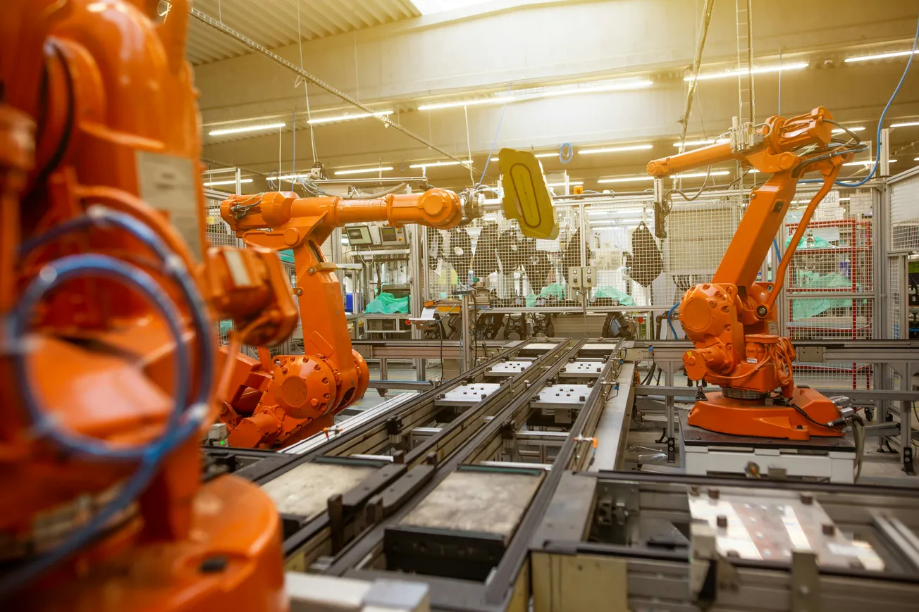 Orange robotic arms work in an industrial factory setting, handling materials on a conveyor system. The scene is bright and organized, conveying efficiency.