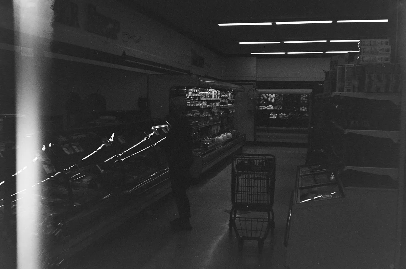 Dimly lit grocery store aisle with blurry figure and shopping cart. Shelves stocked with various goods. Mood is shadowy and mysterious.