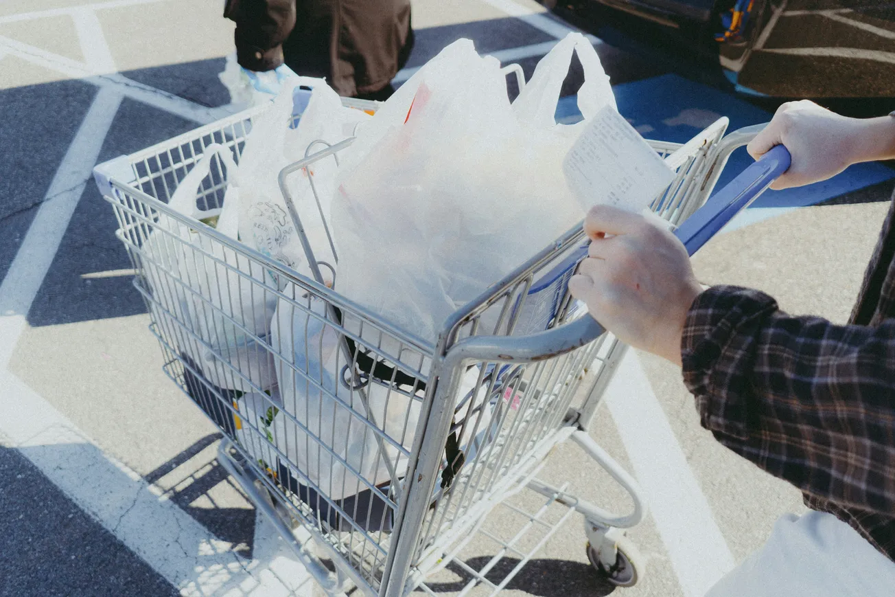 A person pushes a shopping cart filled with plastic bags in a sunny parking lot. They hold a receipt, reflecting a typical grocery shopping scene.