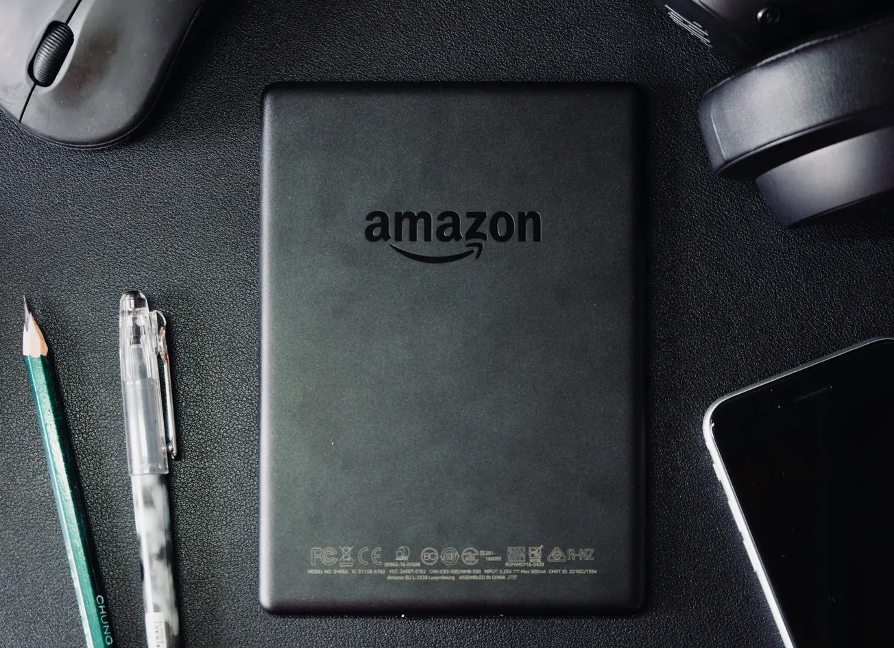 A black Amazon Kindle placed on a dark desk surrounded by a pencil, pen, computer mouse, headphones, and a smartphone, conveying a tech-focused mood.