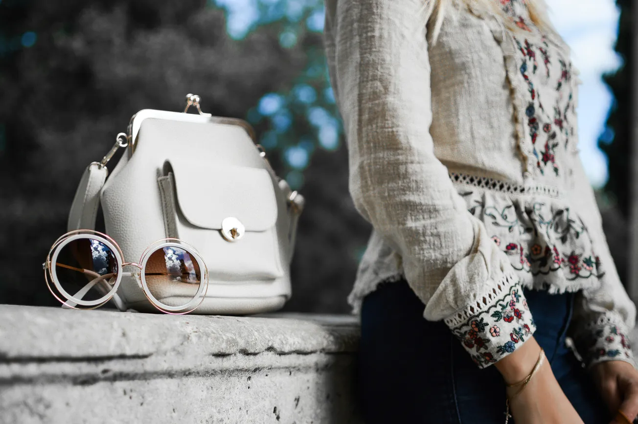 A stylish white bag and round sunglasses rest on a stone ledge next to a person in a patterned blouse, conveying a chic, casual vibe outdoors.