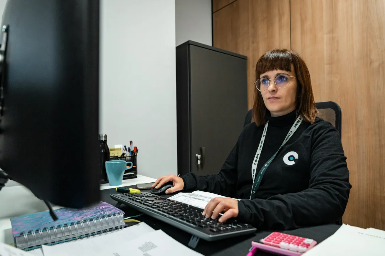 A focused woman wearing glasses and a black sweater works at a computer in an office. A pink calculator and documents are on the desk, with a cabinet behind her.
