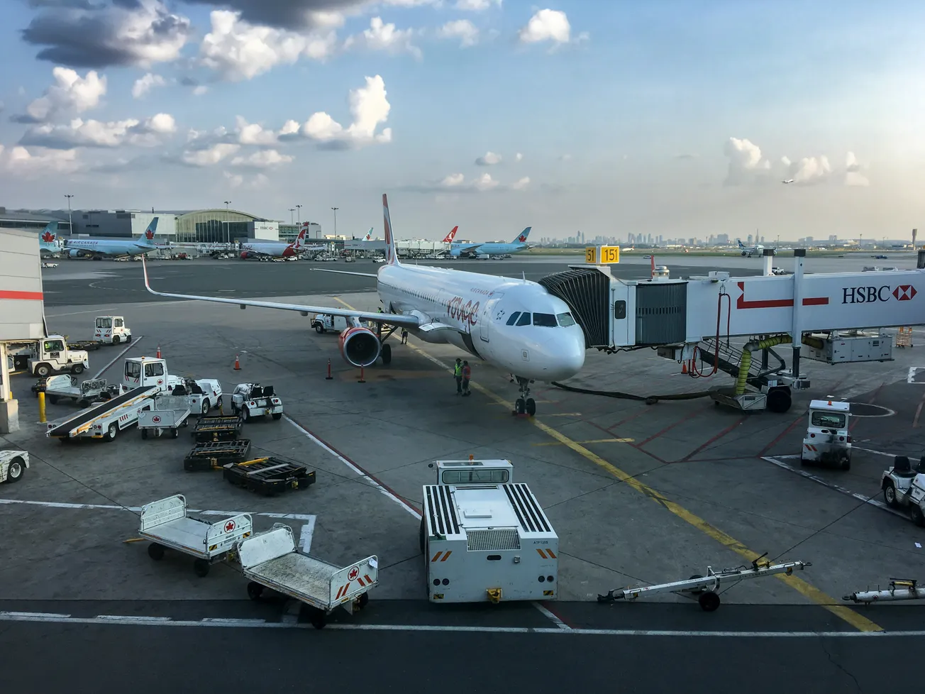Airplane at airport gate with a jet bridge connected. Ground crew and vehicles are busy around the plane under a partly cloudy sky, creating a bustling atmosphere.