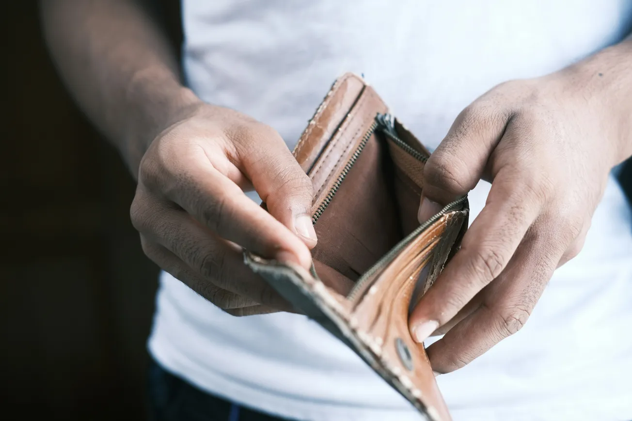 A person wearing a white shirt holds an open brown leather wallet with no cash inside, conveying a sense of financial emptiness and concern.