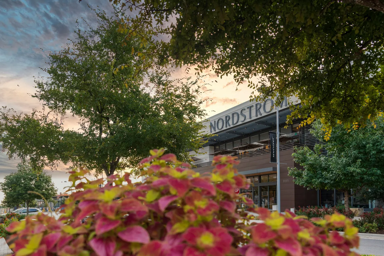 Nordstrom storefront at sunset, framed by lush green trees and vibrant pink flowers in the foreground, creating a serene and inviting atmosphere.