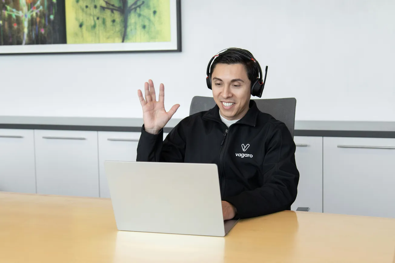 A person wearing a black jacket and headset sits at a table, smiling and waving at a laptop during a video call in a modern, brightly lit office.
