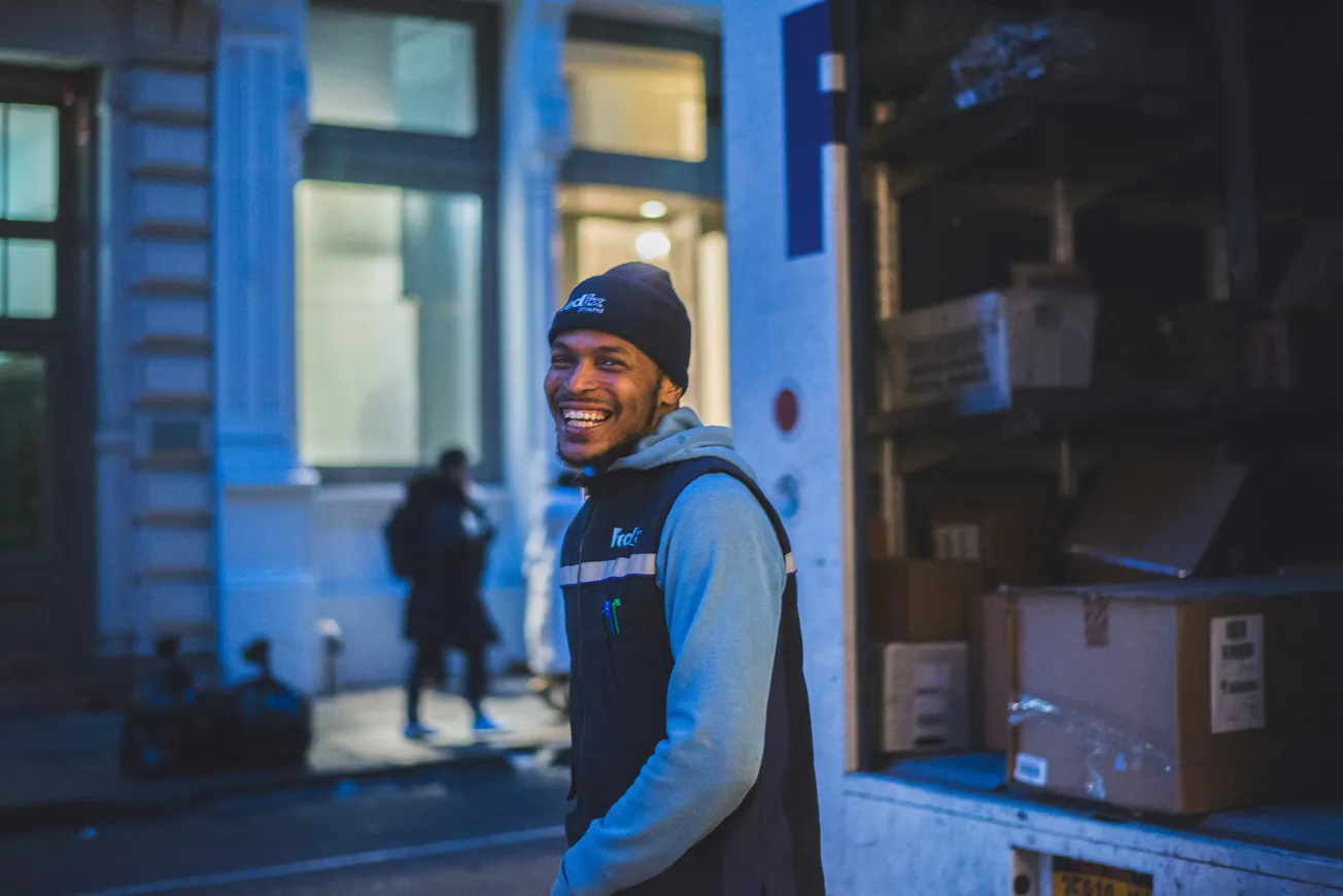 A man in a beanie smiles warmly beside an open delivery truck on a dimly lit street. The scene conveys a sense of joy and camaraderie.