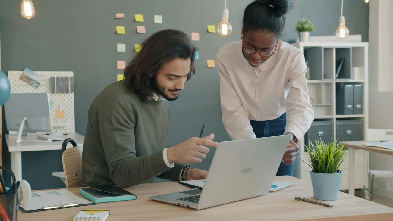 Two colleagues collaborate at a desk with a laptop in a modern office. A wall with colorful sticky notes is visible. The atmosphere is focused and collaborative.