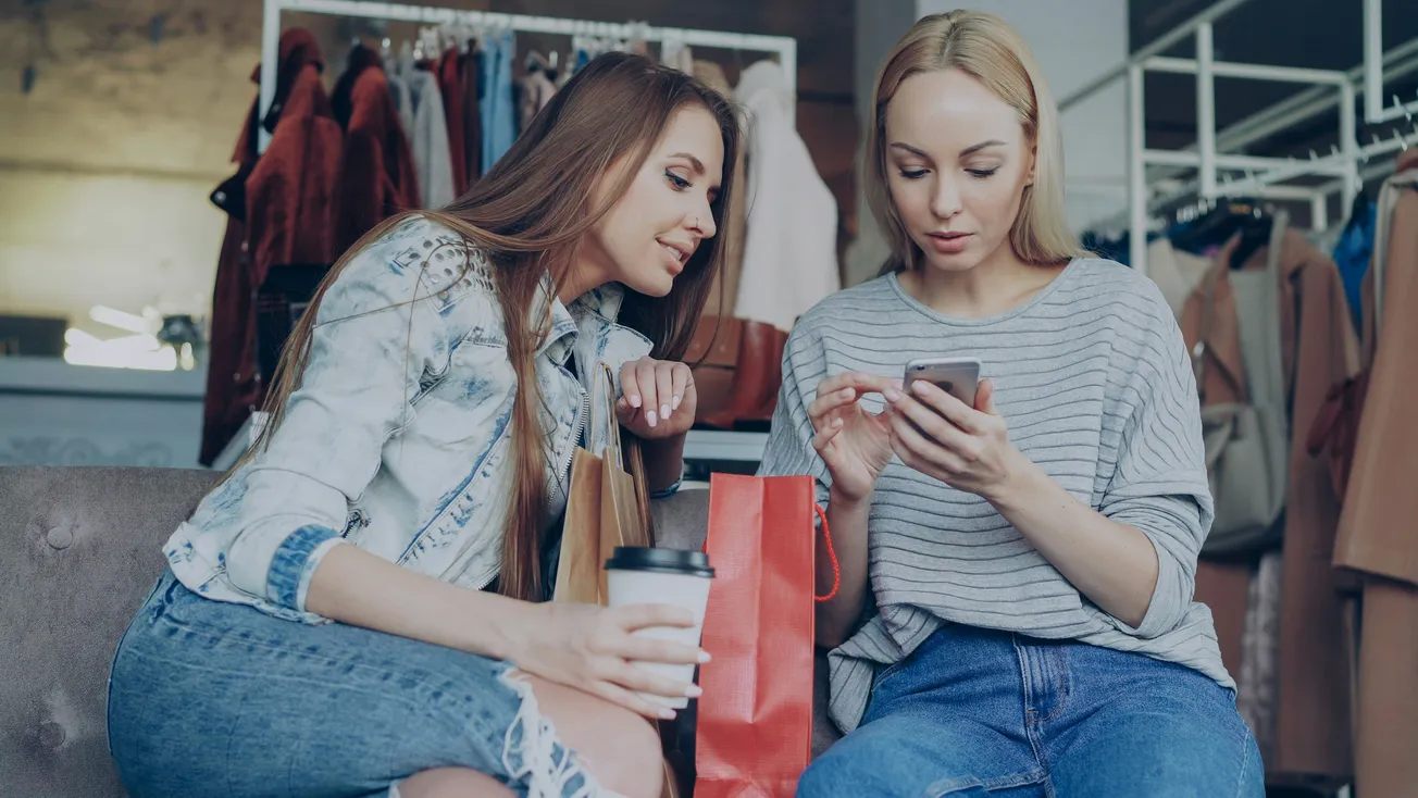 Two women sit on a couch in a clothing store. One wears a denim jacket, holding a coffee cup. The other in a striped top focuses on her phone. Shopping bags nearby.