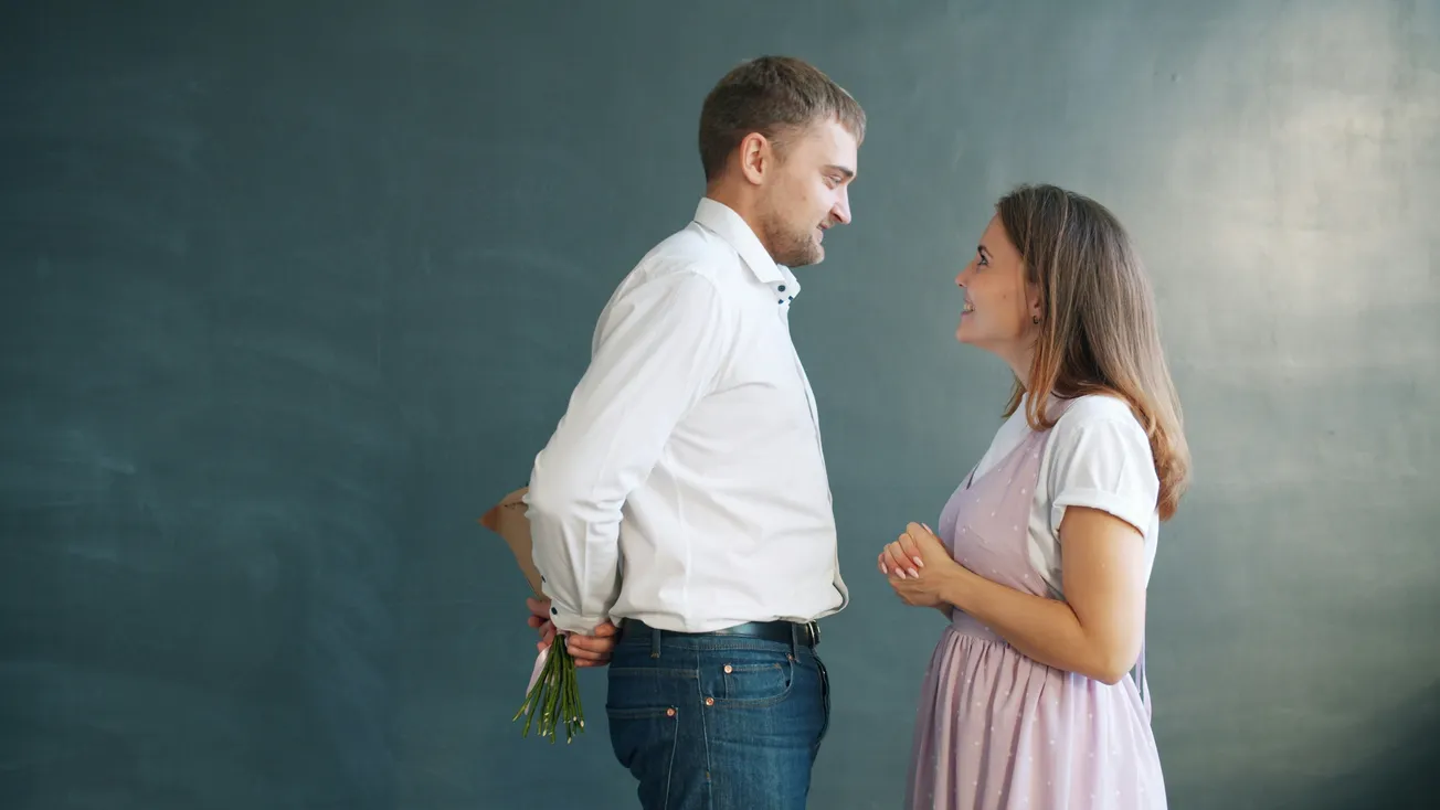 Man in a white shirt smiles at a woman in a lavender dress, holding a bouquet behind his back, against a blue-gray wall, conveying affection.