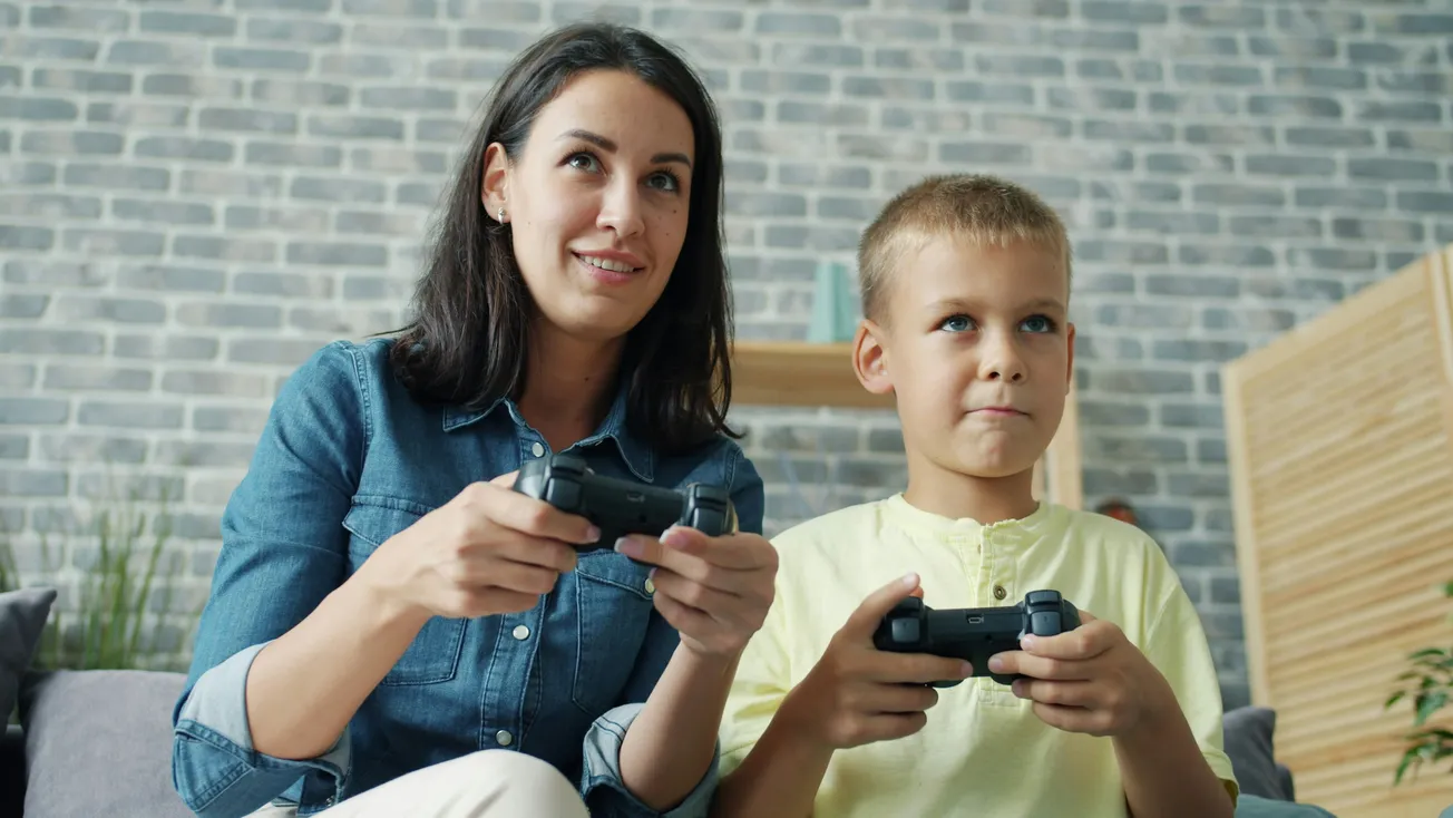 A woman and a boy sit on a couch playing video games with controllers. They appear focused and engaged, sitting against a brick wall backdrop.