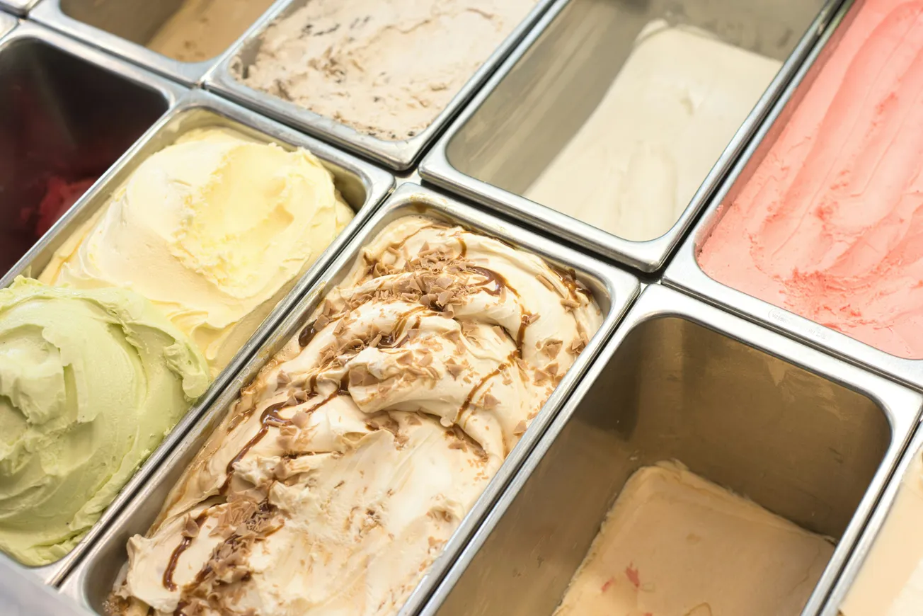 An array of colorful gelato flavors in metal containers, including green, yellow, cream, pink, and swirled caramel. Inviting and delicious display.