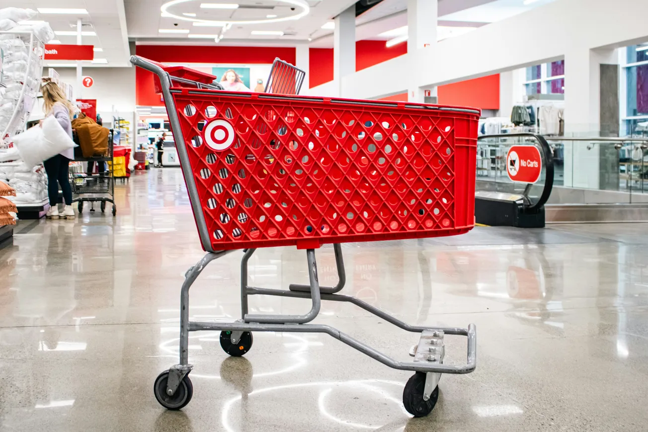 A red shopping cart stands on a polished floor in a department store aisle. Bright lighting and a modern, spacious interior create an inviting atmosphere.