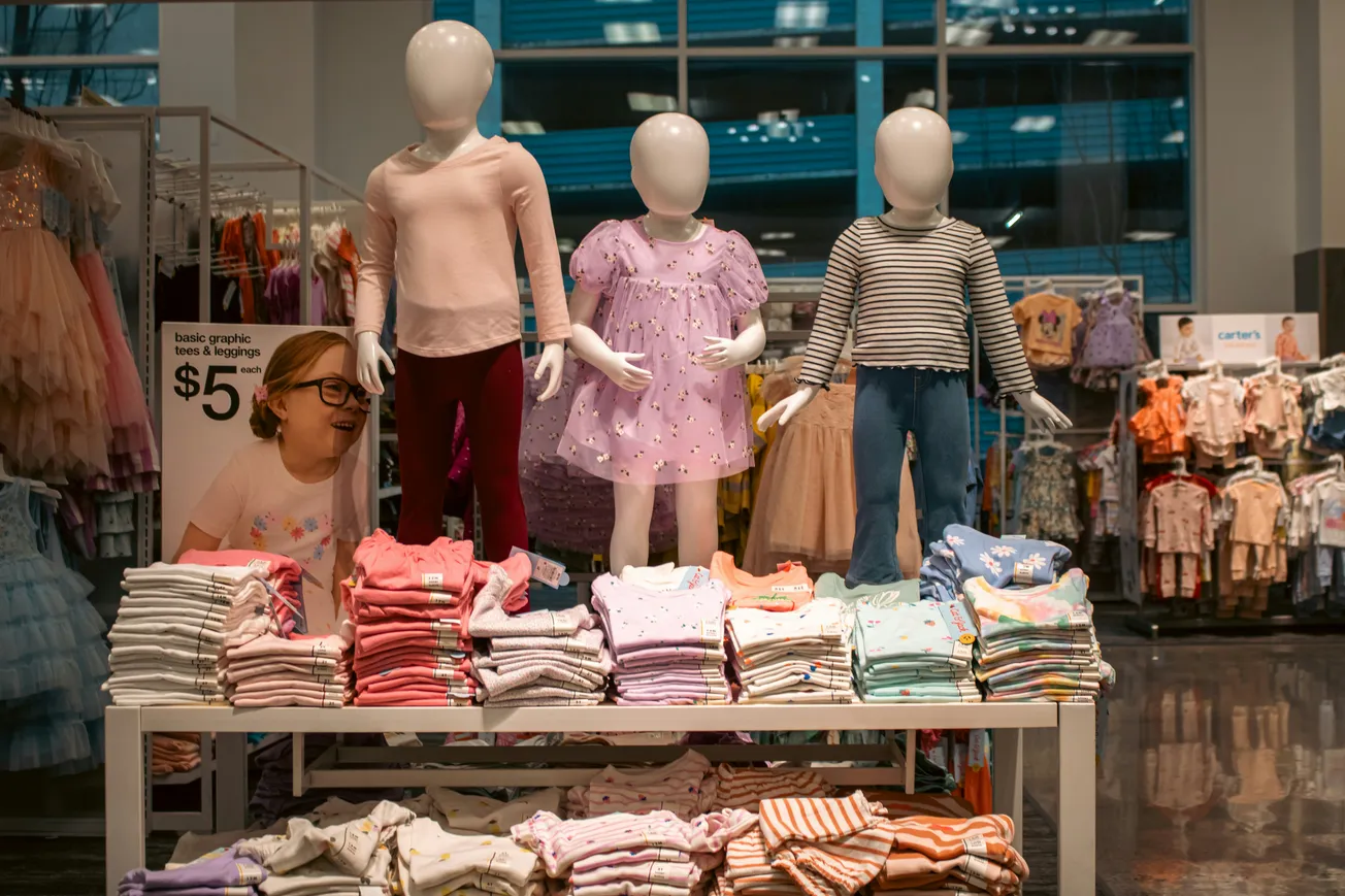 A display in a children's clothing store features three faceless mannequins dressed in pastel and striped outfits. Below are folded clothes, priced at $5.